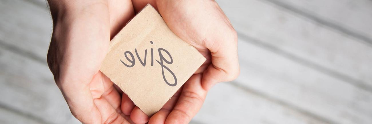 A pair of hands holding a square piece of cardboard with give written on it